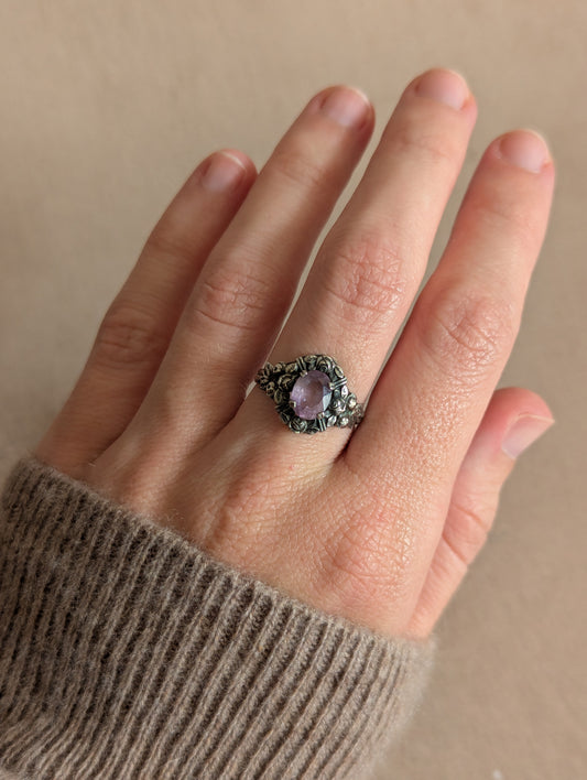 A close-up of a hand wearing a vintage-style silver ring with a lavender-purple oval gemstone and dark oxidized details, set against a neutral beige background.