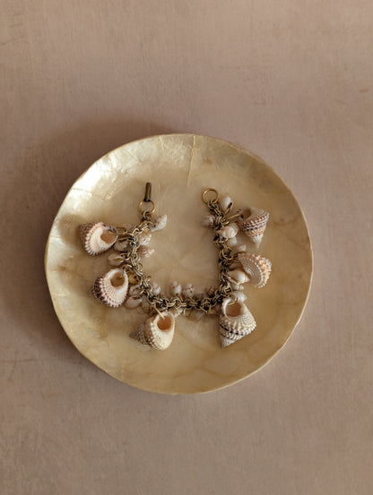 A charm bracelet with various seashells on a chain, displayed on a round, textured cream-colored dish against a neutral beige background.