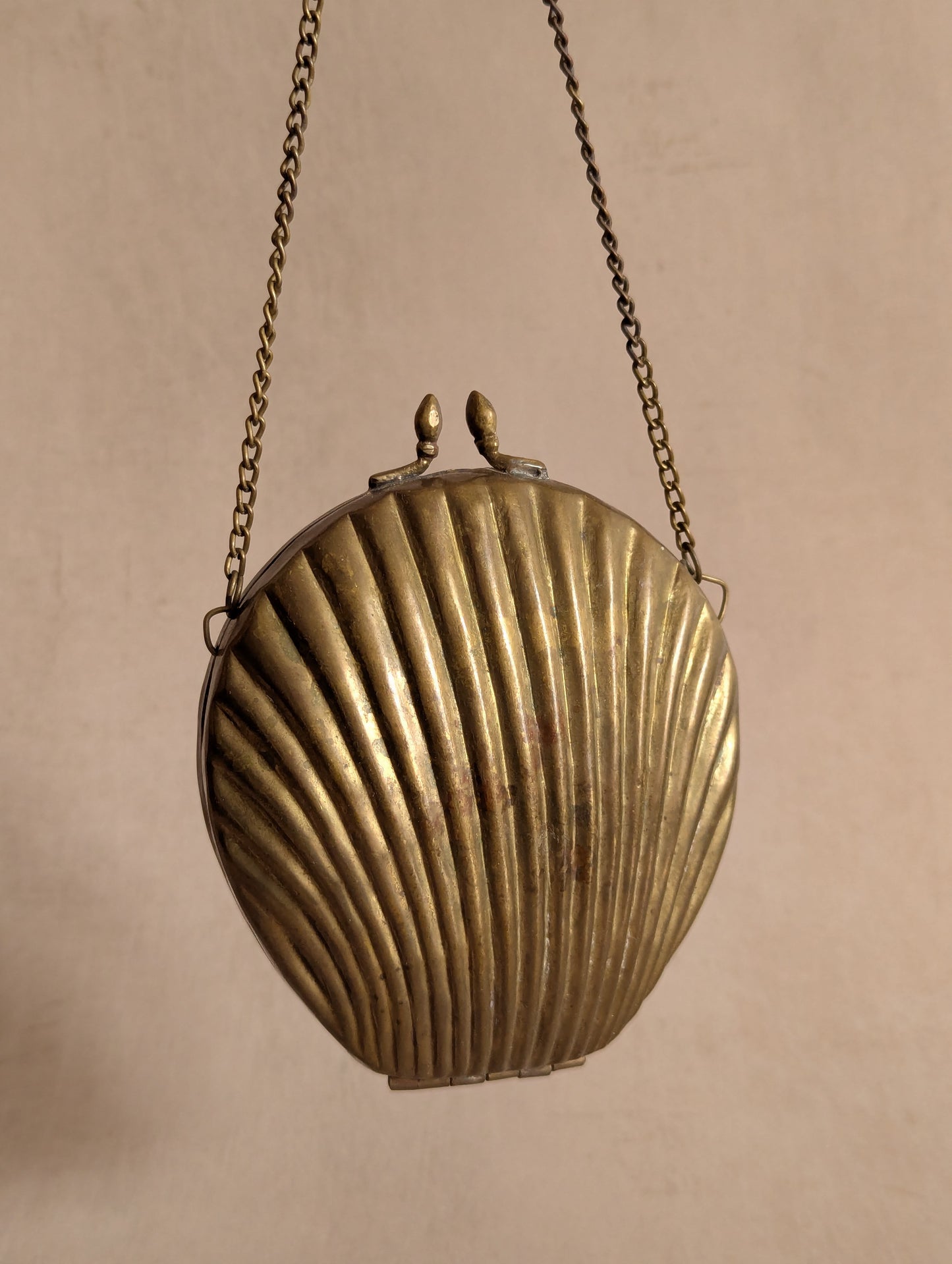 A vintage shell-shaped metal clutch purse with a ribbed seashell design, hanging from a chain against a plain beige background.