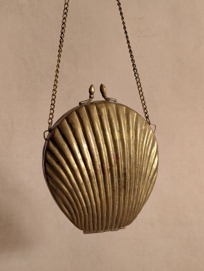 A vintage shell-shaped metal clutch purse with a ribbed seashell design, hanging from a chain against a plain beige background.