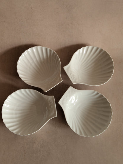 Four white ceramic bowls shaped like scallop shells, arranged in a square pattern on a neutral beige background.