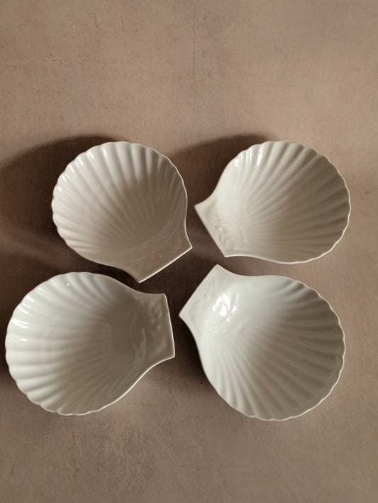 Four white ceramic bowls shaped like scallop shells, arranged in a square pattern on a neutral beige background.