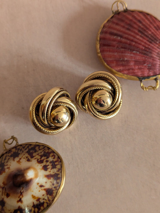 A pair of gold-toned twisted knot earrings with a central dome, displayed on a neutral beige surface alongside decorative seashell-shaped jewelry pieces.