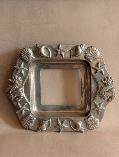 A square silver-toned decorative tray with a raised ocean-themed border featuring starfish, seashells, and small sea creatures, shown on a beige background.