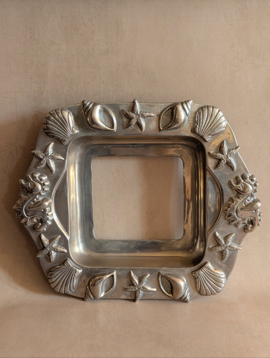 A square silver-toned decorative tray with a raised ocean-themed border featuring starfish, seashells, and small sea creatures, shown on a beige background.