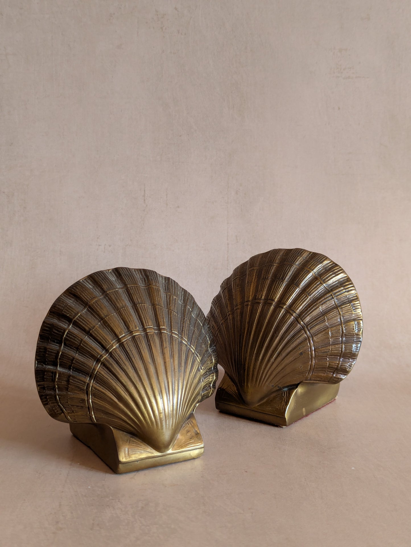 A pair of vintage brass shell bookends shaped like scallop shells, displayed on a neutral beige surface against a textured beige background.
