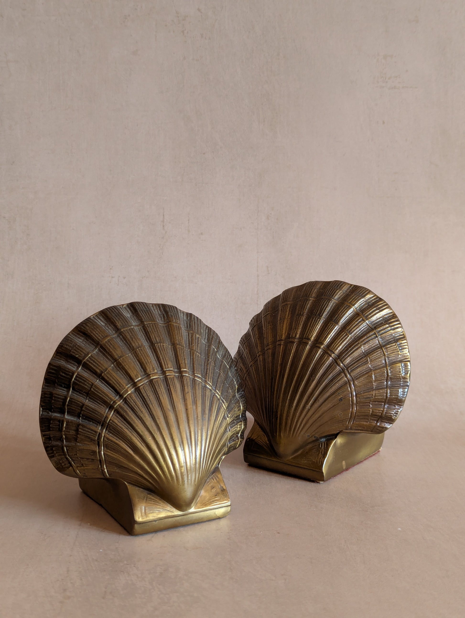 A pair of vintage brass shell bookends shaped like scallop shells, displayed on a neutral beige surface against a textured beige background.