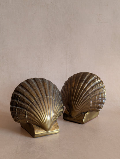 A pair of vintage brass shell bookends shaped like scallop shells, displayed on a neutral beige surface against a textured beige background.