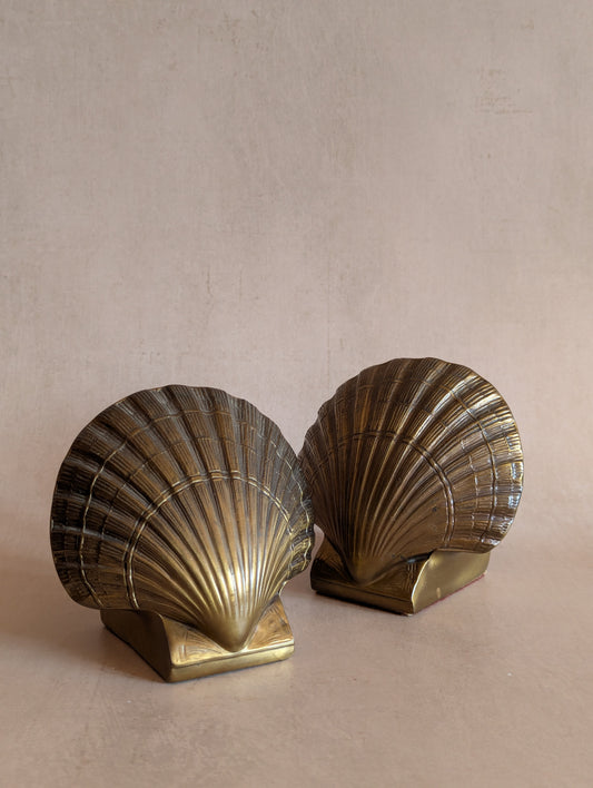 A pair of vintage brass shell bookends shaped like scallop shells, displayed on a neutral beige surface against a textured beige background.